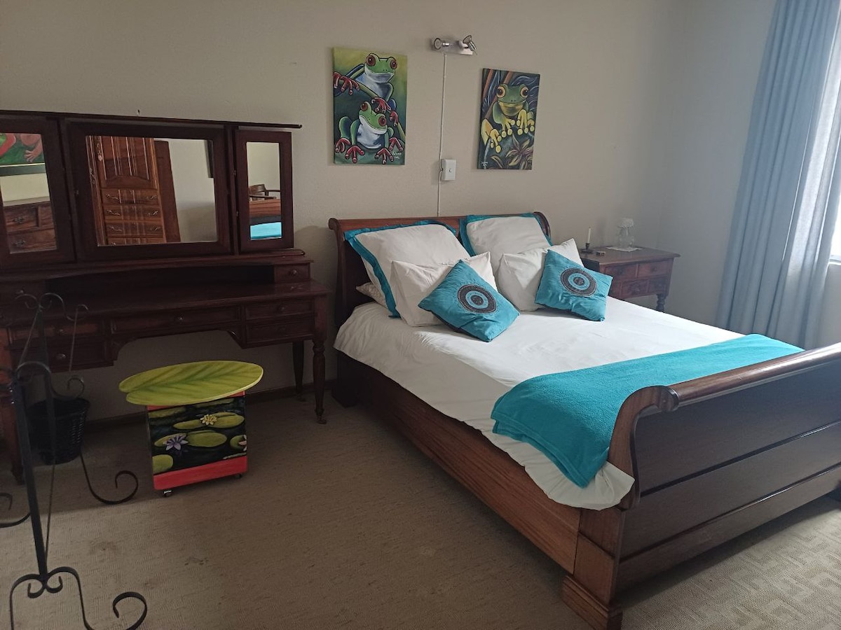 A room is presented with a wooden sleigh bed adorned with white linens and turquoise throw pillows. A matching wooden vanity with a mirror and two bedside tables complement the decor. Colorful artwork is displayed on the walls, while soft natural light enters through a window dressed with light curtains.