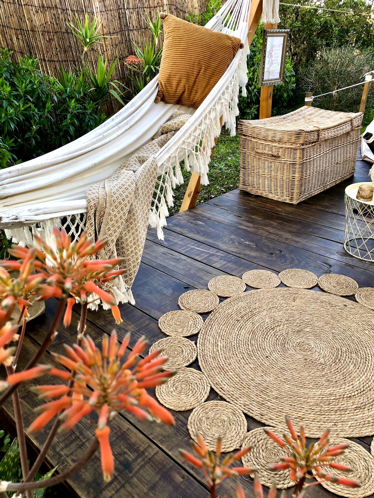 A cozy outdoor relaxation area features a hammock adorned with a decorative pillow, surrounded by greenery. A woven storage basket and a circular natural fiber rug are placed on a wooden deck, complemented by vibrant flowers that add a pop of color to the serene setting.