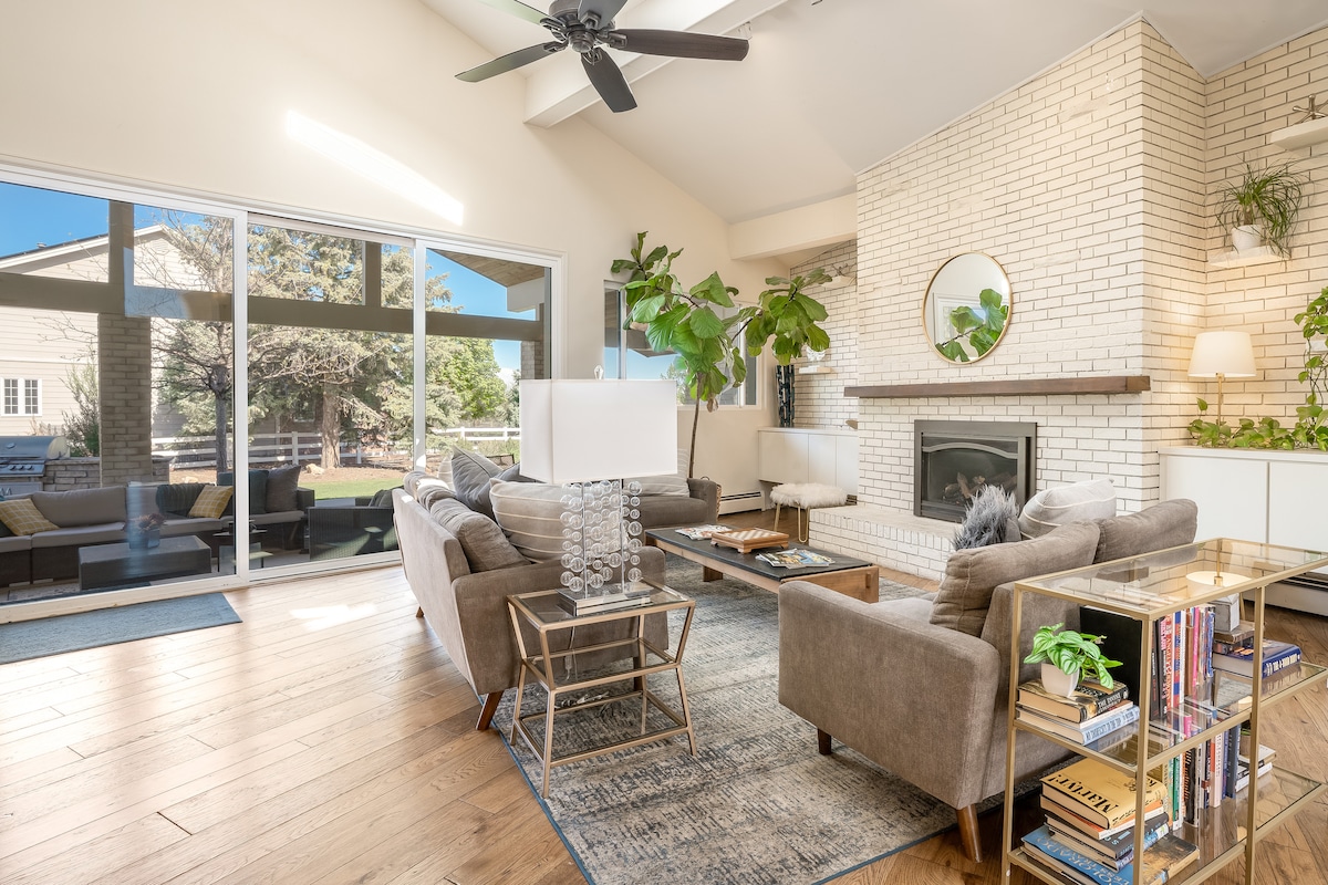 A spacious living area features a comfortable arrangement of sofa seating complemented by two large plants. Natural light floods the room through expansive windows, illuminating the neutral tones and modern decor, including a stylish fireplace and a circular mirror.