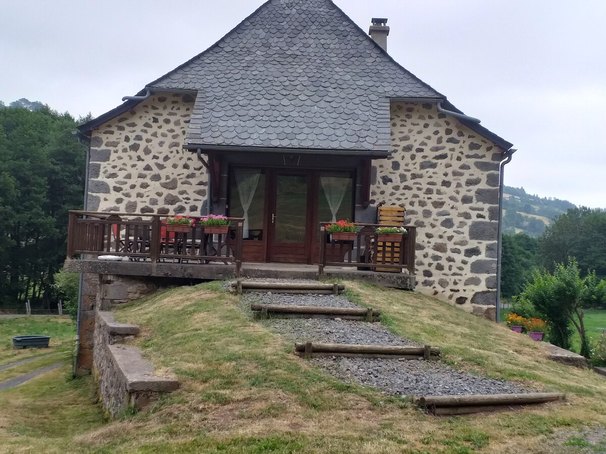 The exterior of the stone building features a sloped grassy area leading to a wooden deck. Flower boxes adorn the railing, and a set of steps provides access to the entrance. A tranquil setting is provided by surrounding trees and landscaping.