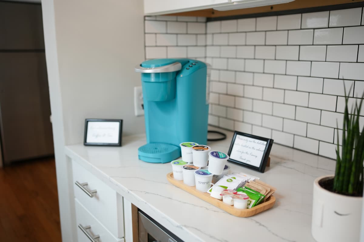 A modern kitchen space features a turquoise coffee maker alongside a selection of coffee pods and creamers arranged on a wooden tray. Framed signs provide information about coffee options. The countertop is complemented by white tile backsplash and sleek cabinetry.