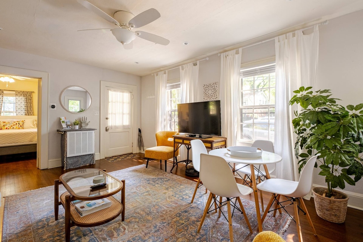 A cozy living space features a comfortable seating area with stylish chairs and a wooden table. A large TV is positioned against the wall, and natural light floods through two windows, complemented by sheer white curtains. A potted plant adds a touch of greenery.