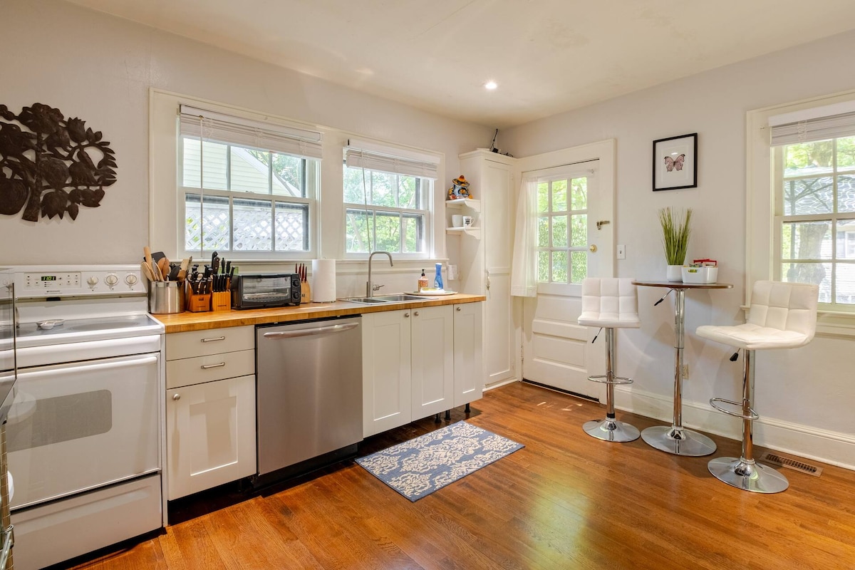 A fully appointed kitchen features modern appliances, including a stove, refrigerator, and microwave. A wooden countertop provides ample workspace, while a bar area with two stools invites casual dining. Natural light fills the space through multiple windows, enhancing the clean, organized layout.