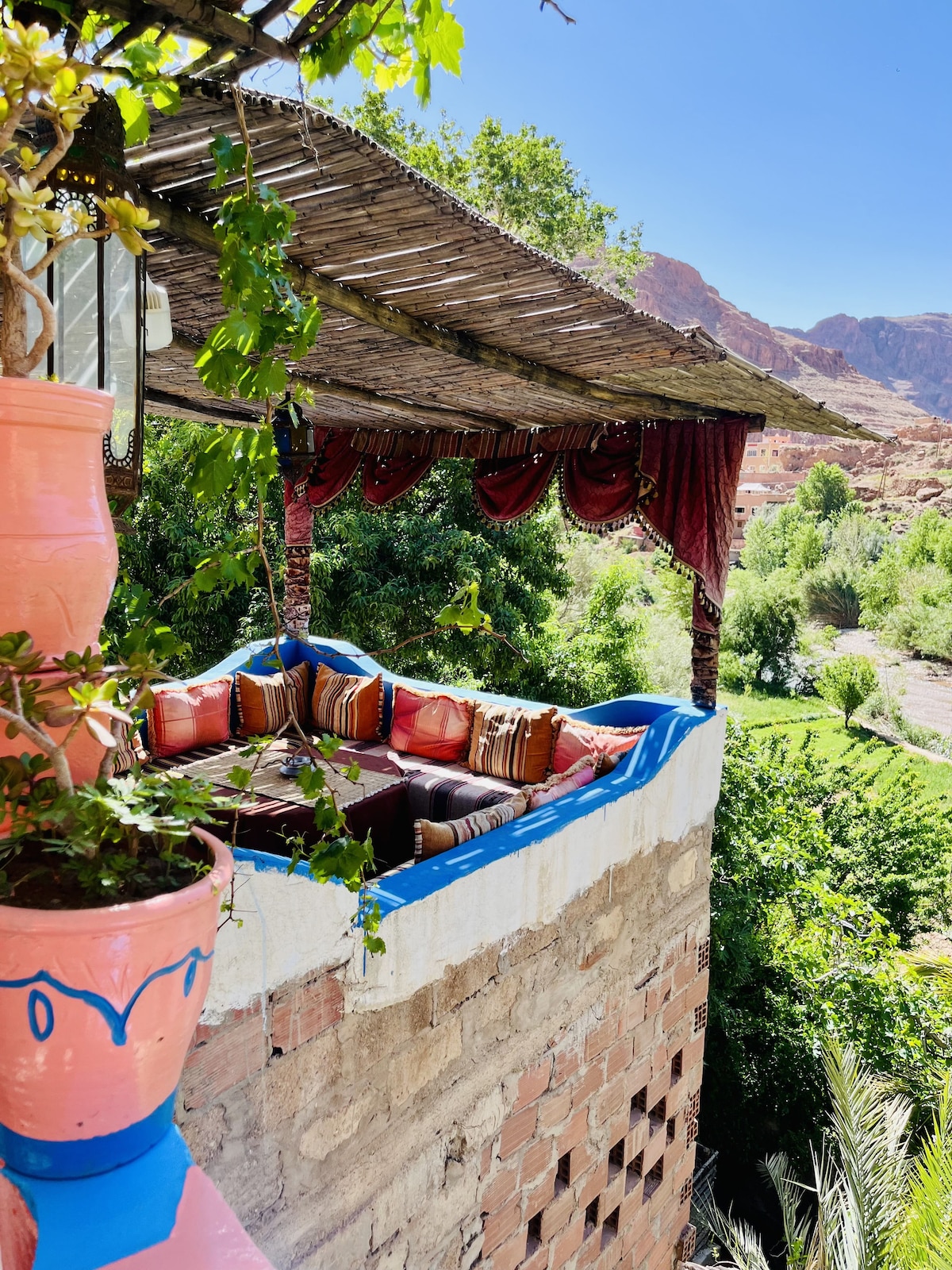 A shaded seating area, adorned with colorful cushions, overlooks lush greenery and mountain views. The structure features rustic brick walls and a thatched roof, with vibrant pots and draped fabric adding to the inviting outdoor space.