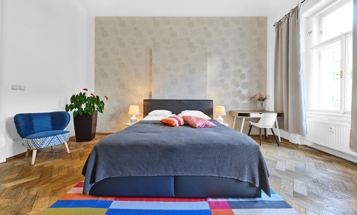 A spacious bedroom is featured with a large bed dressed in dark bedding. A mix of colourful pillows adds a pop of interest. Natural light filters through large windows, and a cozy chair is placed nearby. Elegant wooden flooring enhances the room's ambiance.