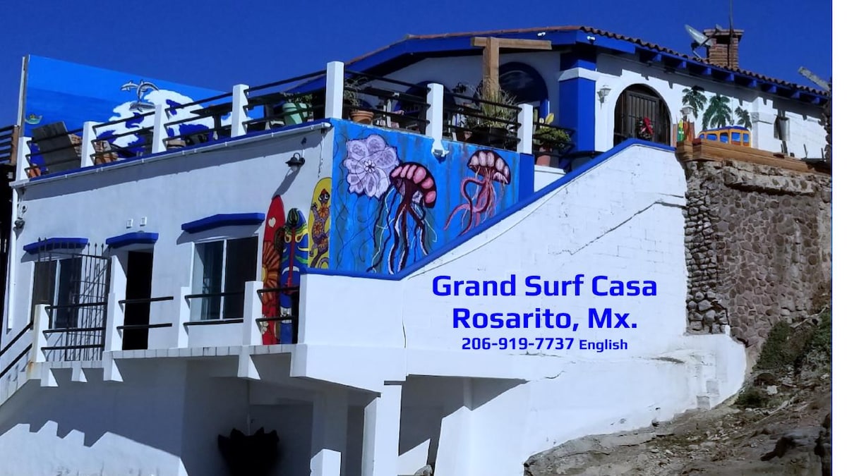 The exterior of Grand Surf Casa is presented, featuring vibrant murals that include colorful jellyfish and local art. The two-story structure showcases a mix of white walls and terracotta accents, complemented by a sweeping staircase leading to the entrance. A clear blue sky overhead enhances the lively atmosphere.
