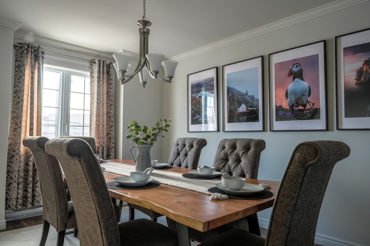 A dining area is presented with a large wooden table surrounded by upholstered chairs. Light streams in from windows adorned with sheer and patterned curtains. Framed artworks featuring scenic landscapes and wildlife decorate the walls, enhancing the room's ambiance.