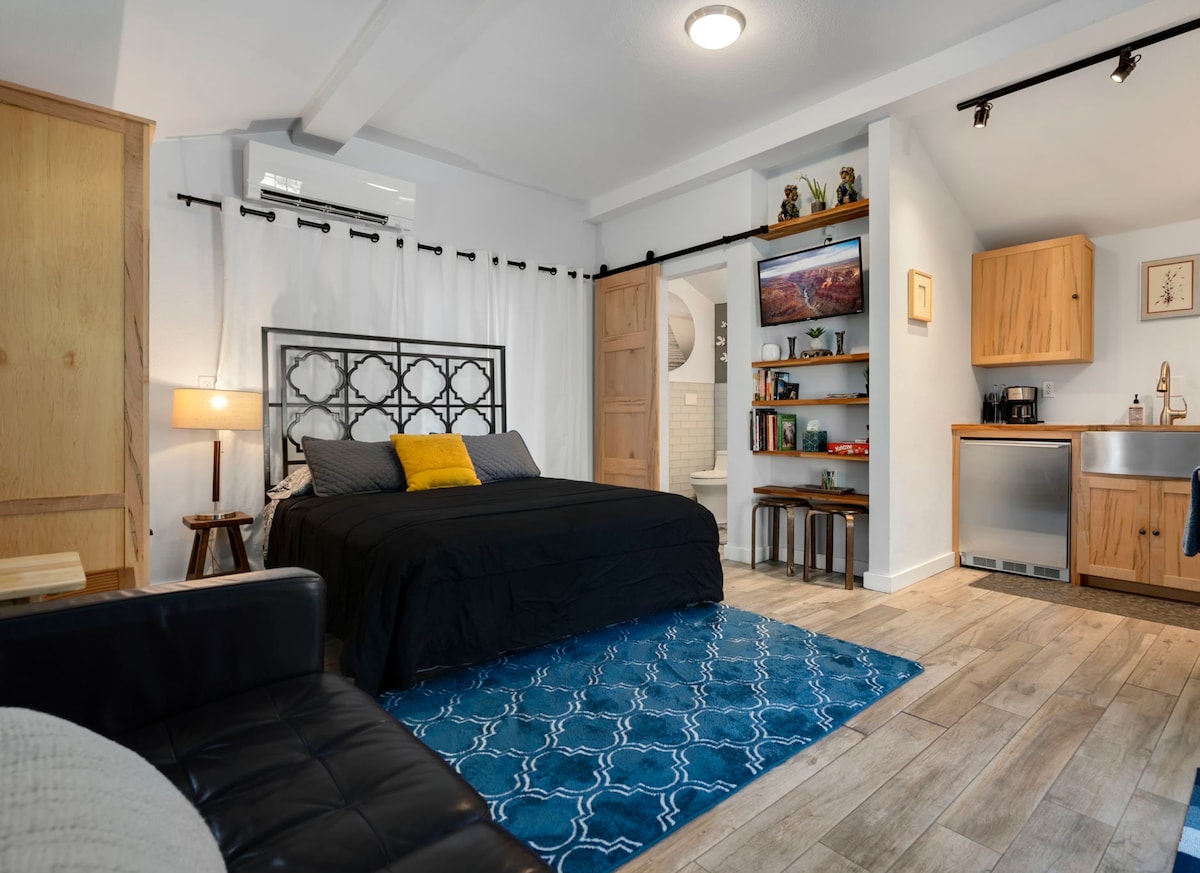 A cozy living space showcases a spacious bed adorned with a black coverlet and yellow accent pillow. A blue area rug anchors the room, complemented by natural wood cabinetry and shelves filled with decor. A kitchenette area is visible, featuring stainless steel appliances.
