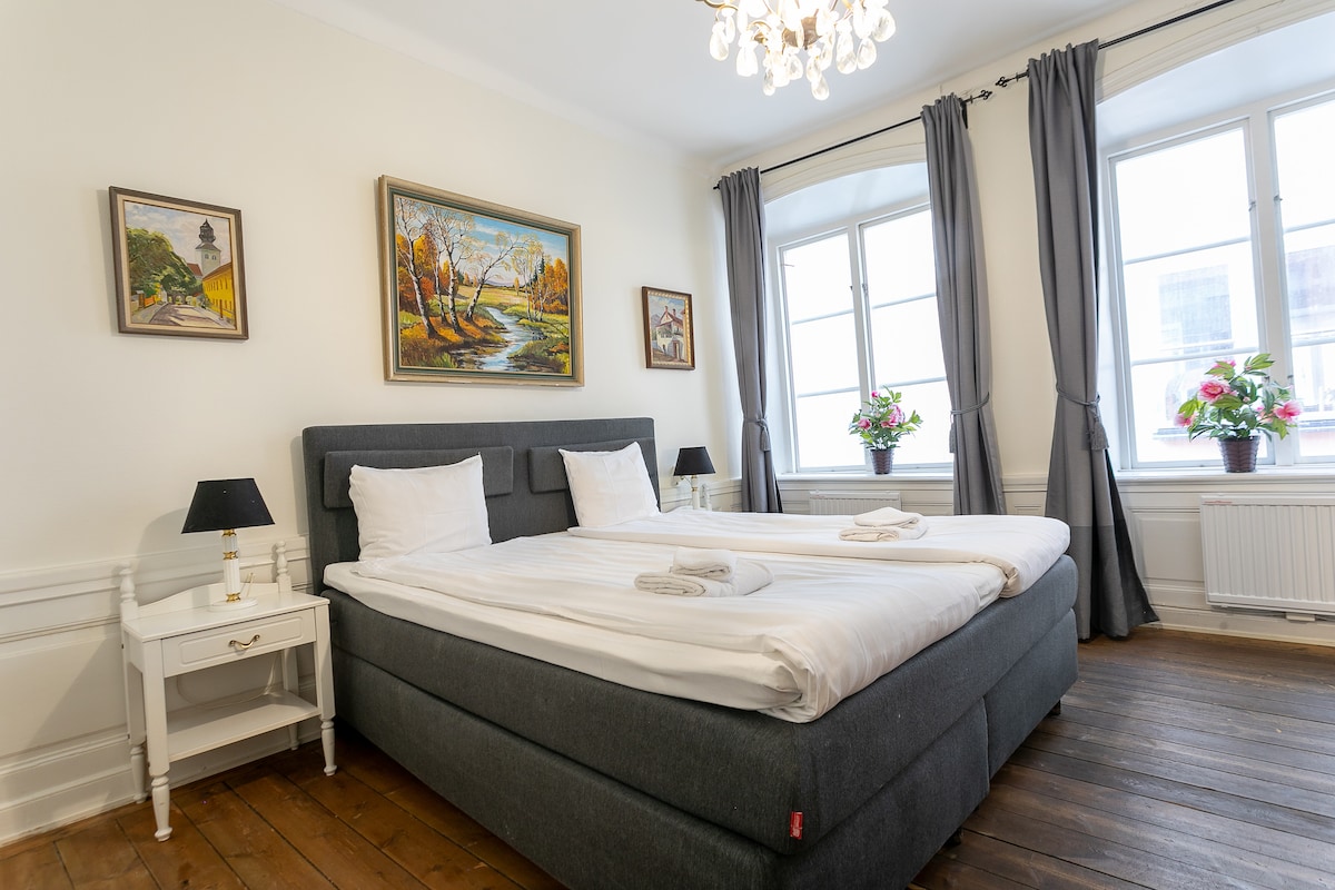 A well-lit bedroom features a spacious bed with two separate mattresses, adorned with fresh white linen. Soft grey curtains frame large windows that provide ample natural light. A floral painting and two bedside lamps enhance the decor, accompanied by a wooden floor that adds warmth to the space.
