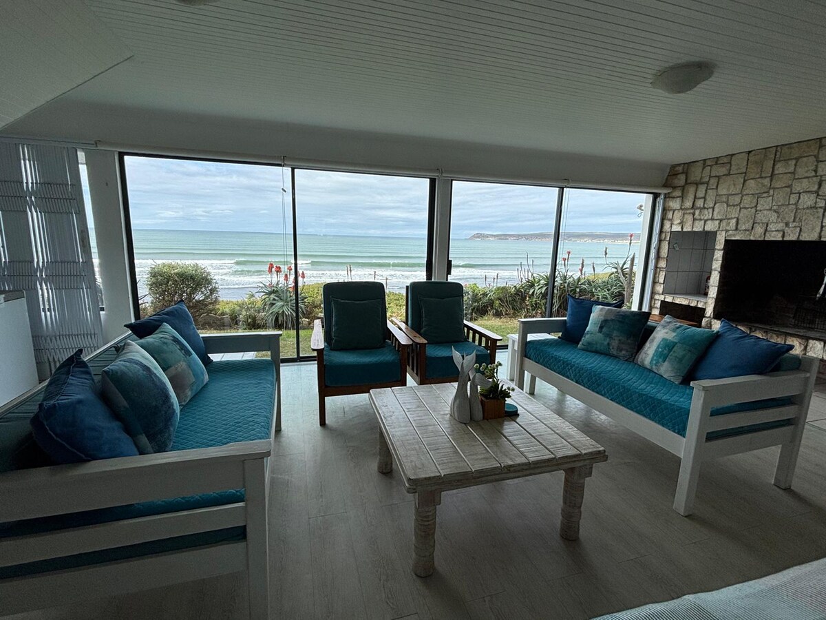 A comfortable living area features large glass windows offering ocean views. Two cushioned armchairs and a sofa adorned with patterned cushions surround a wooden coffee table. Natural light fills the space, highlighting the airy atmosphere and stone accent wall adjacent to the indoor barbecue.