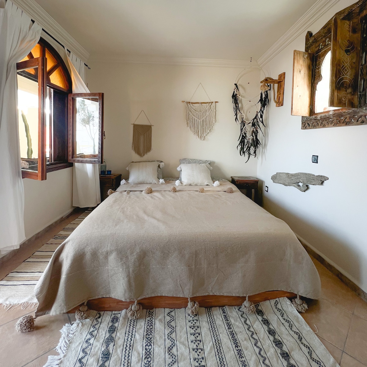 A spacious bedroom features a large bed adorned with a neutral-toned blanket, complemented by light-colored pillows. Natural light enters through two wooden windows, framing views of the outside. Decorative wall hangings and a textured rug enhance the serene atmosphere of the room.