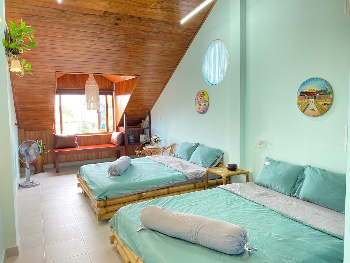 A serene sleeping area is furnished with two spacious beds adorned with teal bedding. Wooden accents and natural lighting from large windows create a calming atmosphere. Decorative wall art and a cozy seating nook beside the window contribute to the inviting space.