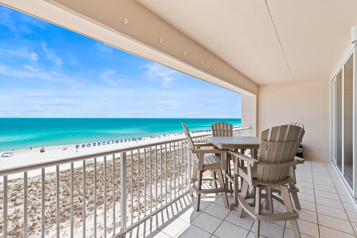 A spacious outdoor patio is furnished with heavy-duty furniture, offering a dining table and chairs. Expansive views of the beach and ocean, showcasing clear turquoise waters and sandy shores, are visible through the large openings. Bright sunlight fills the space, enhancing the coastal ambiance.