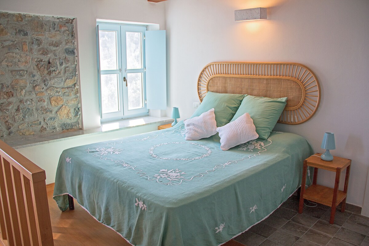 A cozy bedroom features a spacious bed with light blue bedding, complemented by decorative pillows. Natural light enters through a window with blue shutters, and the room is adorned with natural stone accents on one wall. Two bedside tables with lamps provide a soft glow.