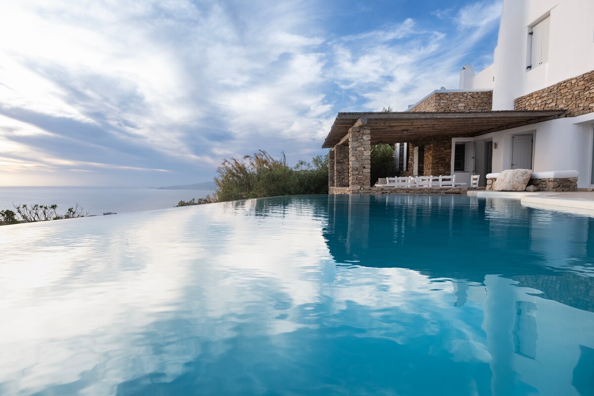 An infinity pool reflects the cloud-streaked sky, extending seamlessly towards the horizon. A covered patio provides shaded seating, with white lounge chairs positioned near the water's edge, inviting relaxation while enjoying coastal views.