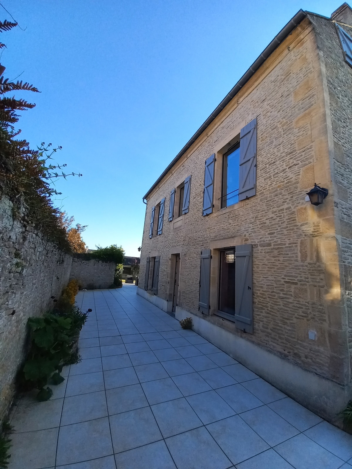 The exterior of the house is displayed, featuring a light stone façade complemented by window shutters. A tiled pathway leads alongside the building, bordered by greenery and stone walls, under a clear blue sky.