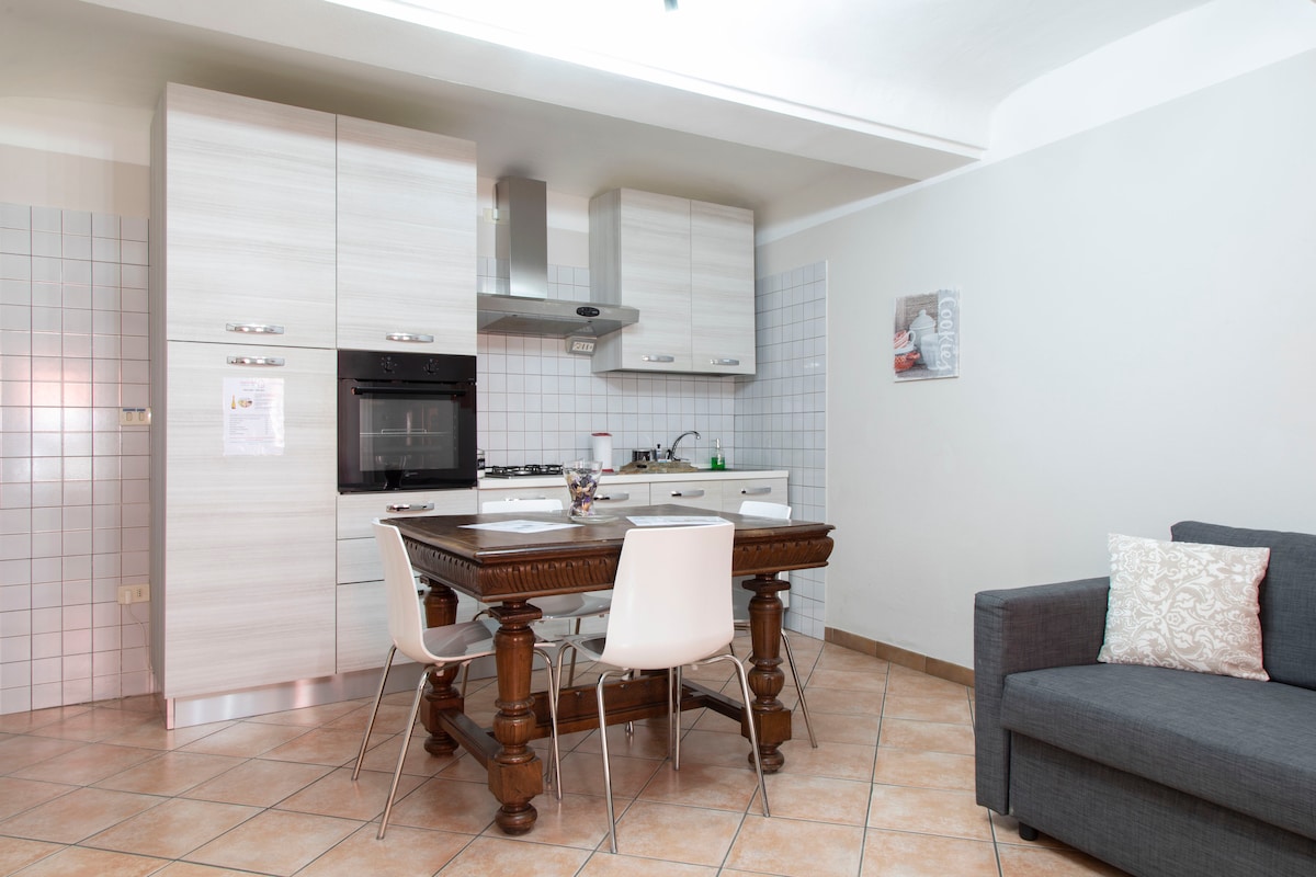 An open-plan kitchen and living area features light-colored cabinetry and a dark wooden dining table surrounded by four chairs. A gray sofa is visible against one wall, creating a functional yet cozy space. A small window allows natural light into the room.