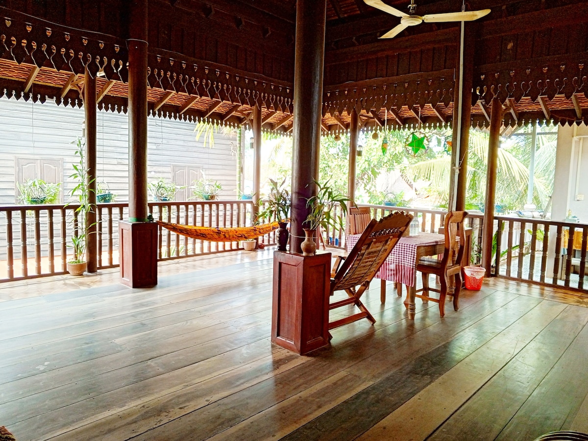 A spacious balcony features wooden flooring and is surrounded by decorative plants. Comfortable seating options include a simple wooden dining table with chairs and a hammock for relaxation. Ceiling fans offer a cooling breeze, enhancing the open-air experience beneath a traditional wooden roof structure.