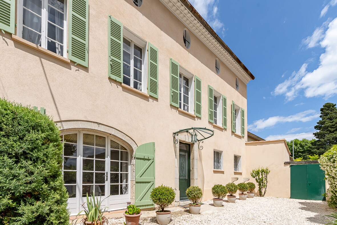 The façade of a Provençal house is presented, featuring light-colored walls with green shutters framing the windows. A covered entrance is highlighted by a decorative awning, while potted plants add greenery to the gravel path. The sky is partly cloudy, creating a bright atmosphere.