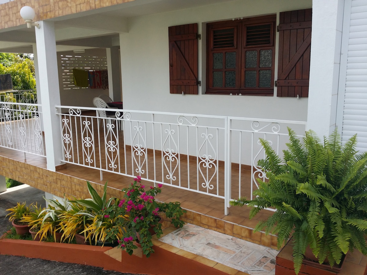 A front porch area is displayed with a decorative white railing. Potted plants, including vibrant flowers and ferns, add greenery. The wooden window shutters are closed, and a cozy seating area is visible in the background, contributing to a tranquil outdoor space.