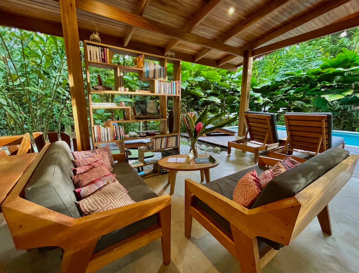 A shared lounge area features wooden furniture, including two armchairs and a comfortable sofa adorned with patterned cushions. A wooden bookshelf holds a selection of books, while lush greenery is visible in the background. Natural light brightens the space.