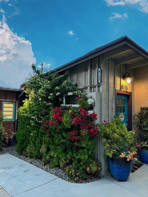 A charming cottage exterior is surrounded by vibrant greenery and blooming flowers. The front door, painted in a welcoming blue, is framed by well-maintained plants in large blue pots. Soft lighting above the door adds a comforting touch to the inviting entrance.