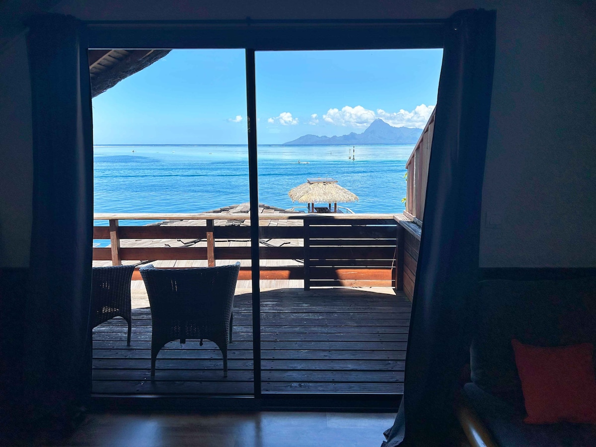 A spacious deck is visible through large sliding glass doors, offering a stunning view of the calm blue lagoon. Two wicker chairs are positioned on the deck, with a thatched umbrella partially seen in the distance, framed by the sky and water.