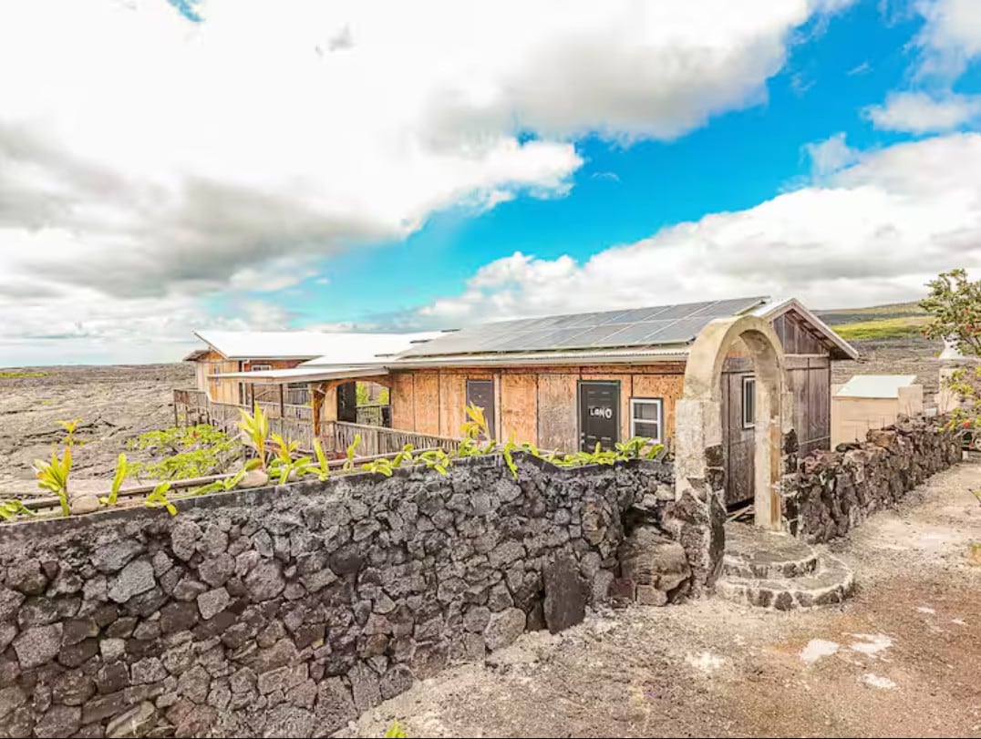 The exterior of a rustic building is showcased against a backdrop of cloudy skies and expansive lava fields. A stone wall with green foliage outlines the entrance, while solar panels are visible on the roof, indicating sustainable energy use.