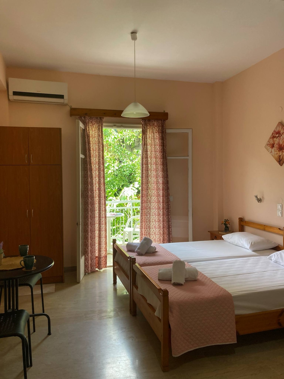 A cozy studio features two single beds dressed in white linens, complemented by a light pink bedspread. A wooden wardrobe and a small dining table with chairs are present. Natural light flows in from the balcony door, framed by patterned curtains that add warmth to the space.