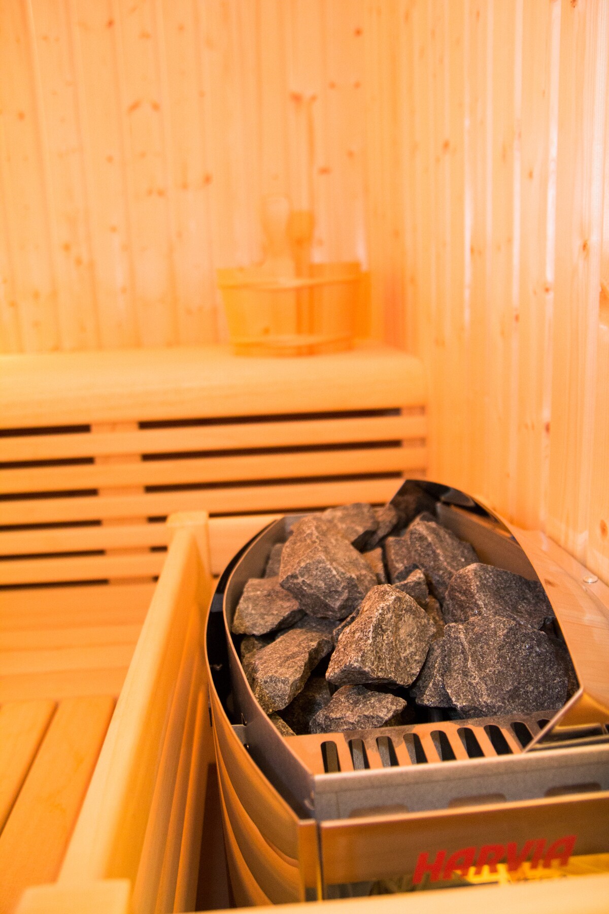 A wooden sauna interior is showcased, featuring a stacked stone heater at the center with gray rocks resting atop. Light wood slats line the walls and benches, complemented by a wooden bucket and ladle placed on a shelf. A warm glow enhances the calming environment.