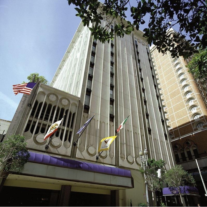 The exterior view of the Donatello Hotel features a multi-story facade with distinct architectural lines. Flags representing the United States and other nations are visible. The entrance is marked by a purple awning and is surrounded by landscaped greenery.