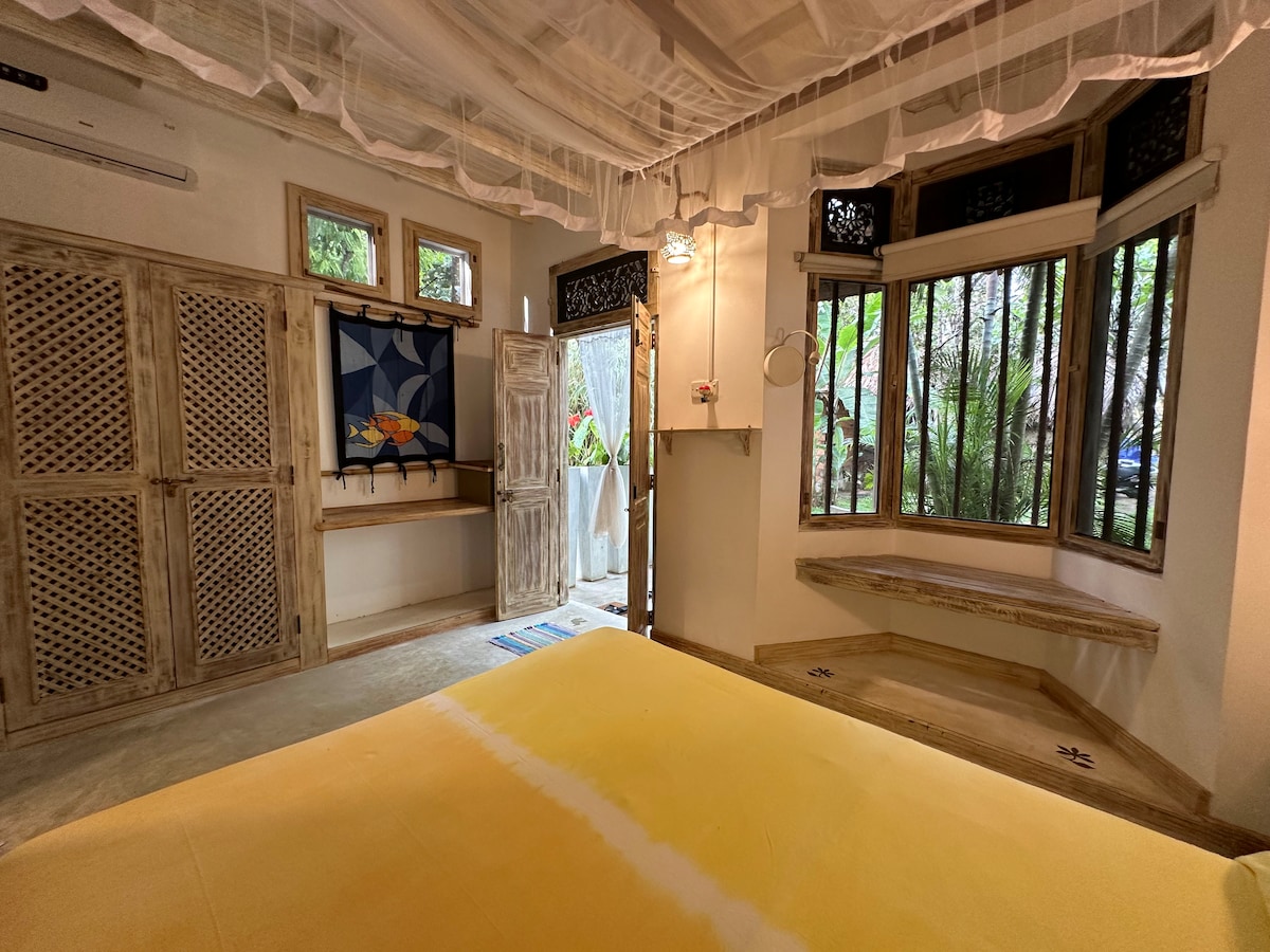 The spacious bedroom features a large yellow bedspread. Natural light enters through multiple windows, highlighting wooden accents and details. A door leads to an outdoor area, with foliage visible through the glass. Decorative elements include a patterned wall hanging and a built-in bench beneath the windows.