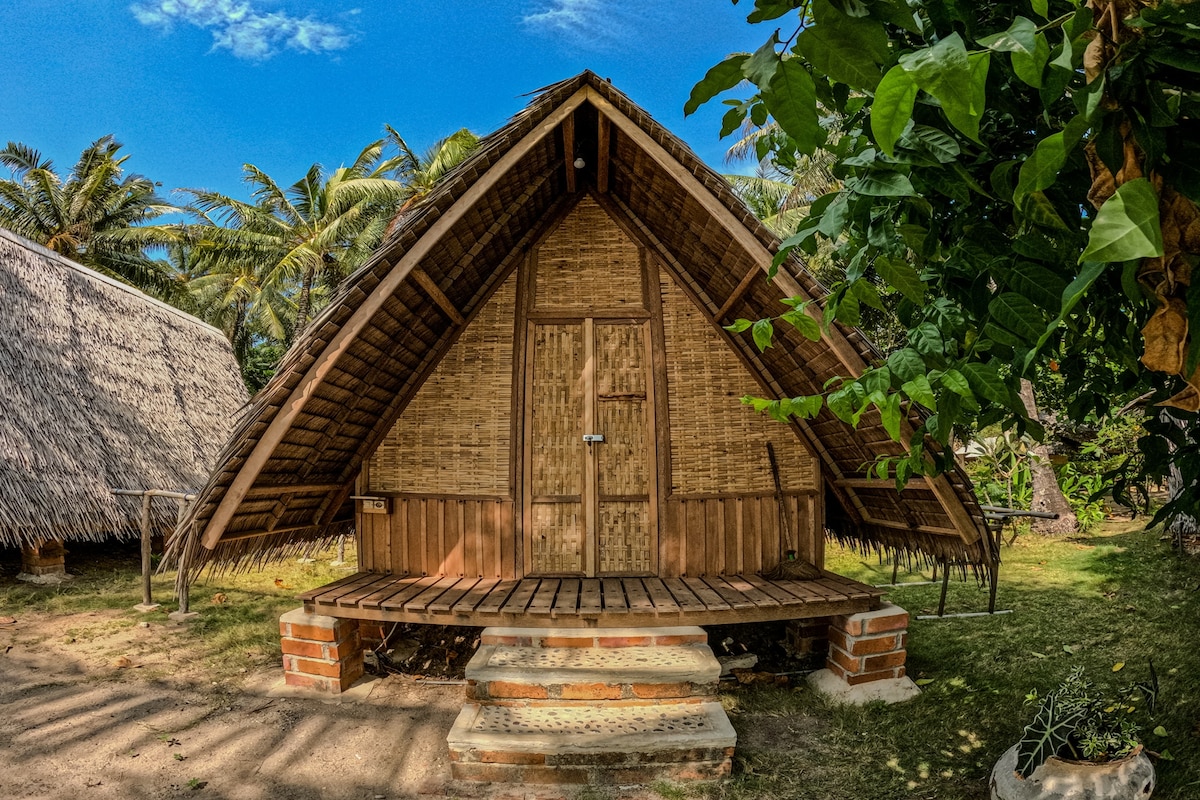 A traditional bungalow features a thatched roof and wooden walls, set amidst lush greenery. Steps lead to a wooden deck in front, providing an outdoor space to enjoy the surrounding nature. The bright blue sky and palm trees enhance the tranquil setting.