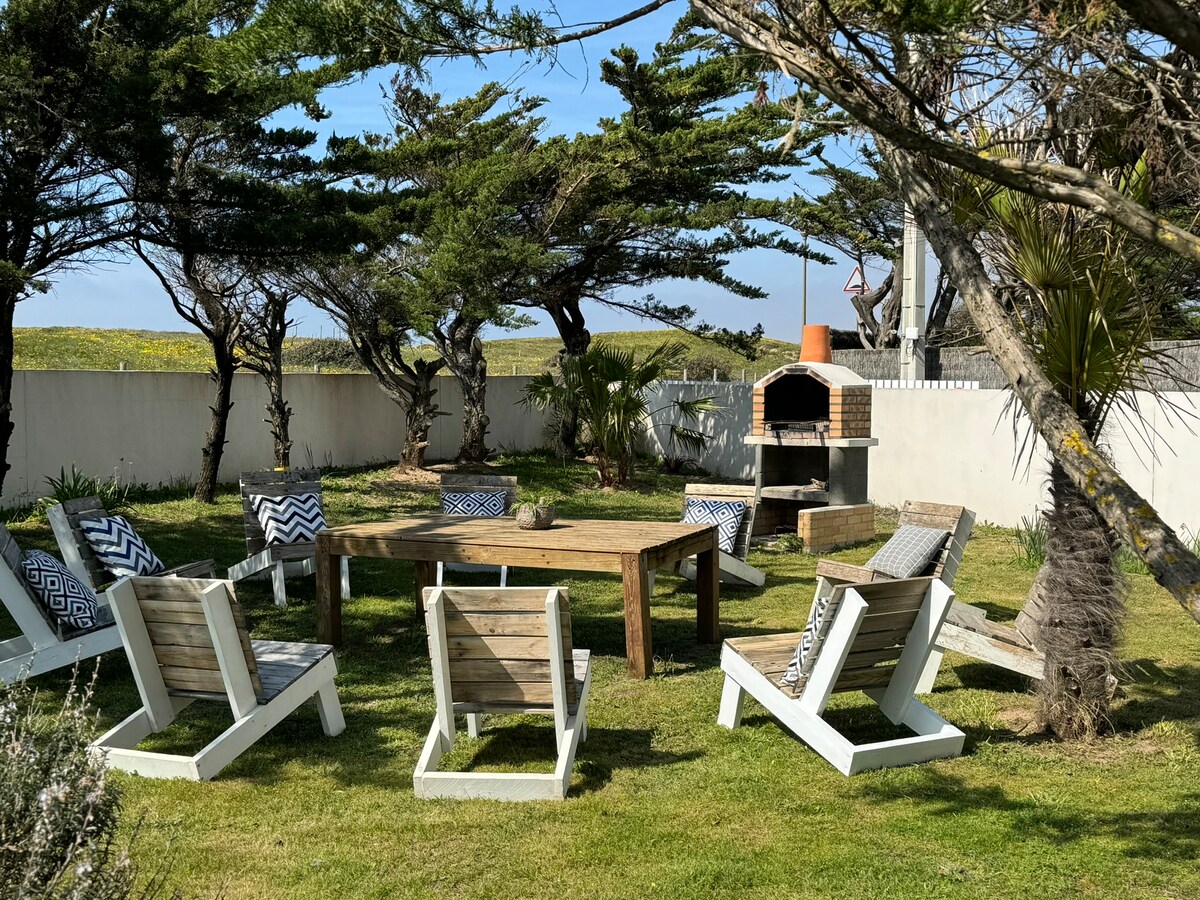An outdoor space features a wooden table surrounded by several lounge chairs, creating a relaxing atmosphere. A stone barbecue is positioned nearby, with tall trees providing shade. Lush green grass covers the ground, enhancing the inviting setting for gatherings.