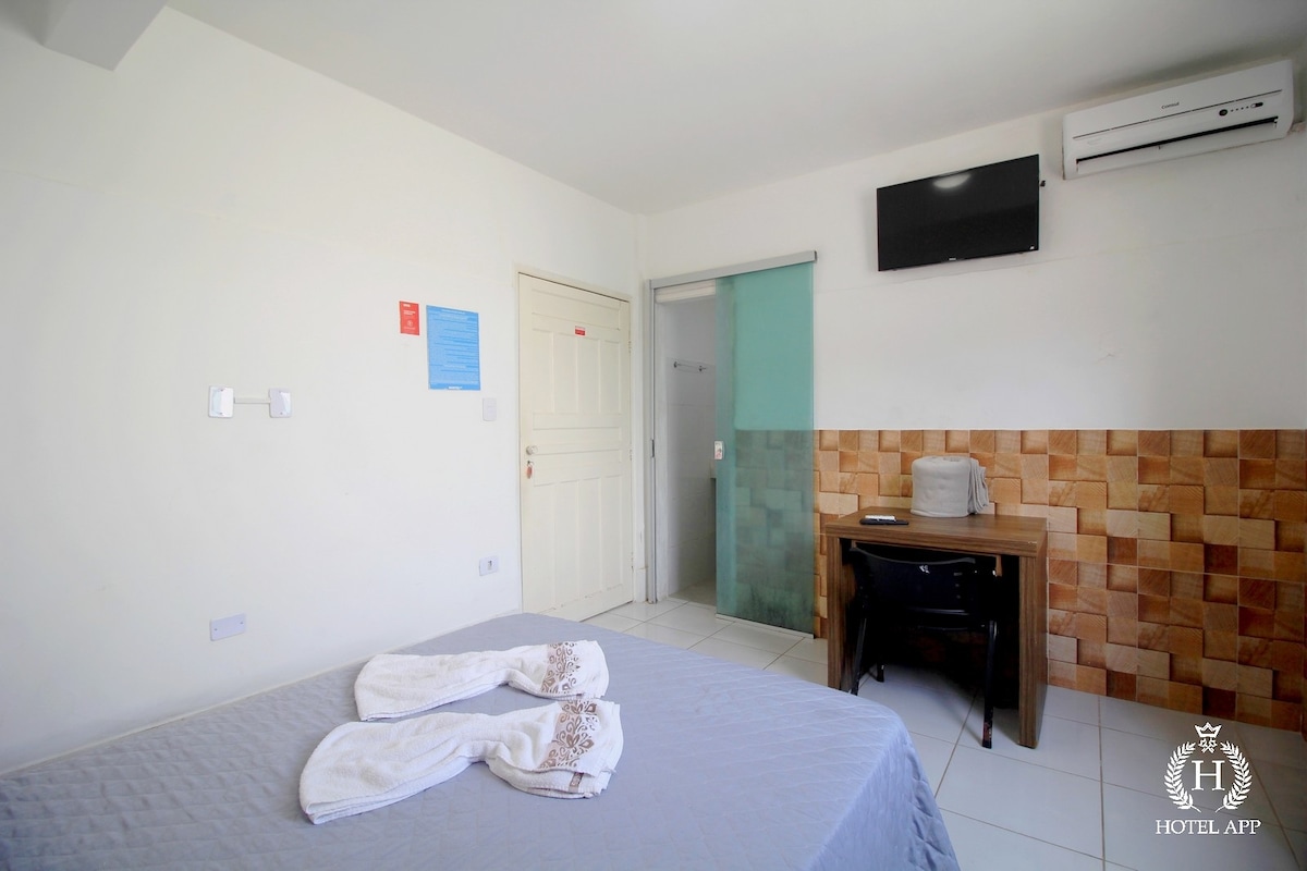 A simple guestroom features a spacious bed with a light-colored bedspread. A wooden desk is positioned nearby, and a flat-screen TV is mounted on the wall. An air conditioning unit is installed above the bed, while light enters through nearby windows.