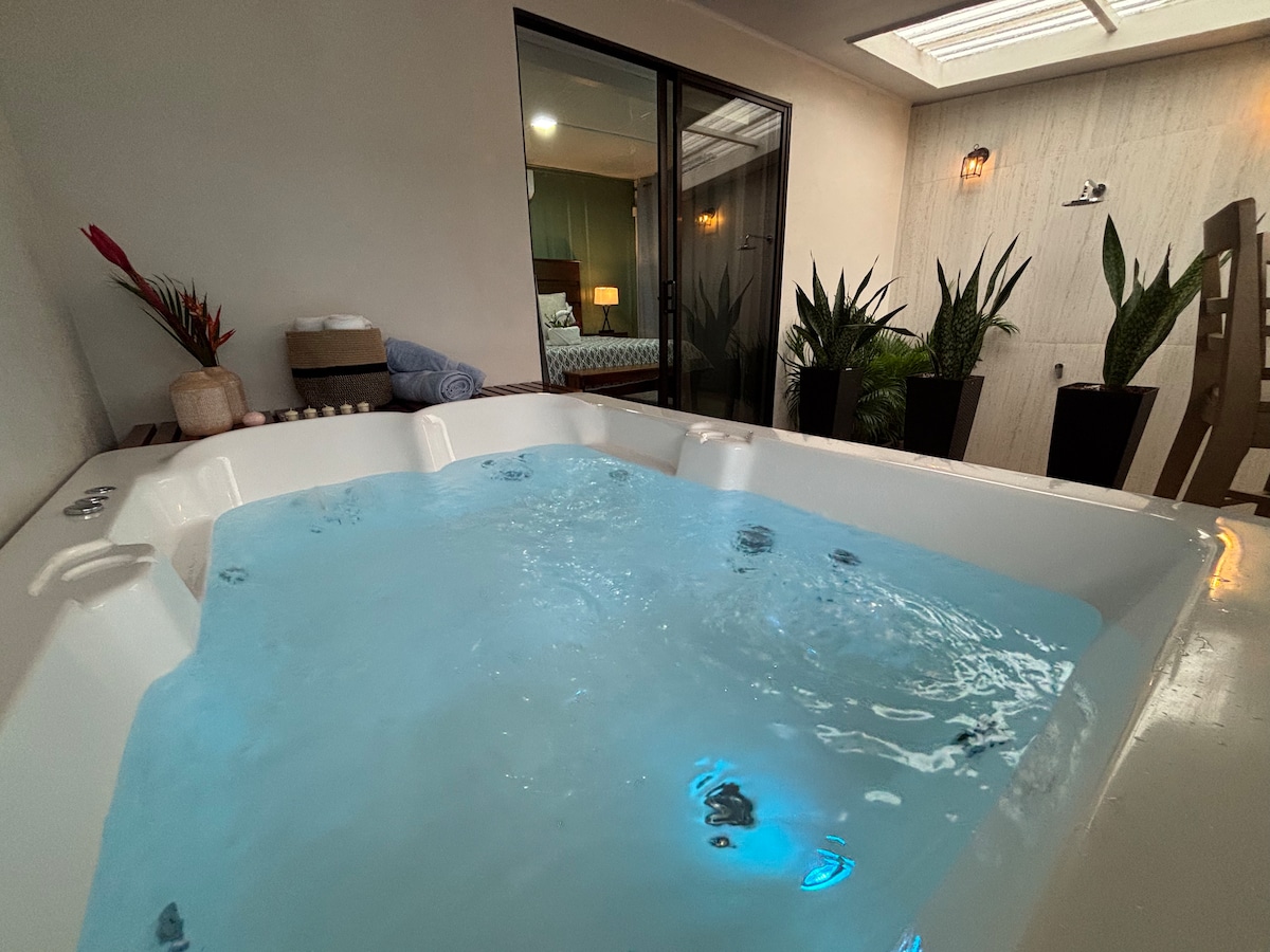 A private hot tub with clear, bubbling water is positioned inside a cozy nook, surrounded by green plants in large pots. Soft lighting is provided by fixtures above, enhancing the tranquil setting that invites relaxation.