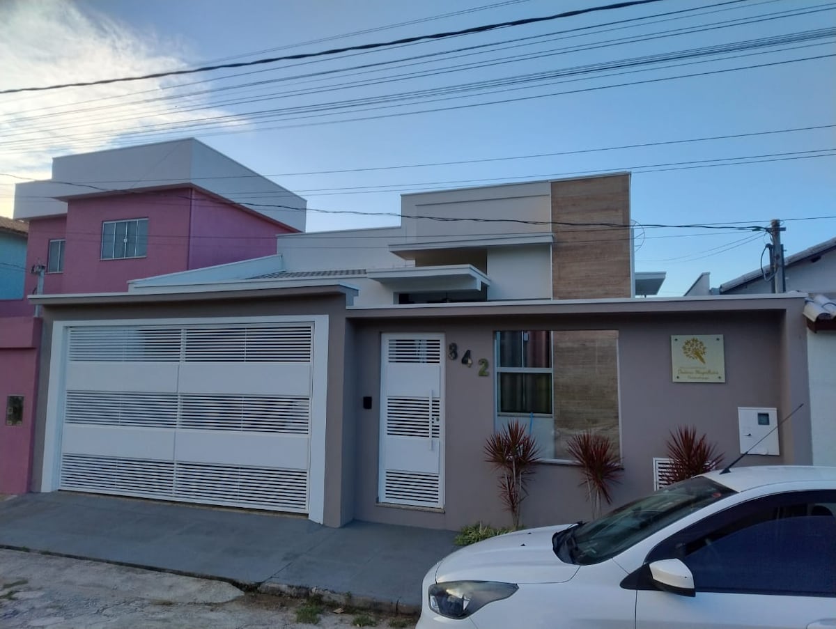 The exterior of a modern house features a sleek design with a mix of light-colored walls and natural stone accents. A spacious driveway leads to a large garage door. Bright landscaping, including decorative plants, enhances the front area. The house is situated in a well-lit neighborhood.