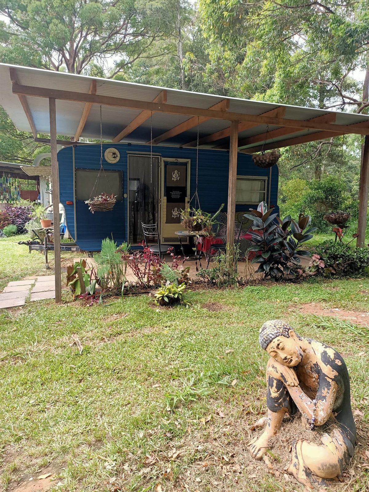 A vintage blue van with a welcoming porch is nestled among lush greenery. The covered area features hanging plants and outdoor seating, while a decorative statue adds an artistic touch to the vibrant garden landscape.