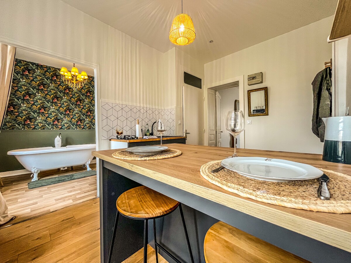 A dining area is displayed with a wooden table and two stools, featuring woven placemats. In the background, a well-equipped kitchen is visible alongside a freestanding bathtub, elegantly positioned against a leafy wallpaper, illuminated by soft overhead lighting.