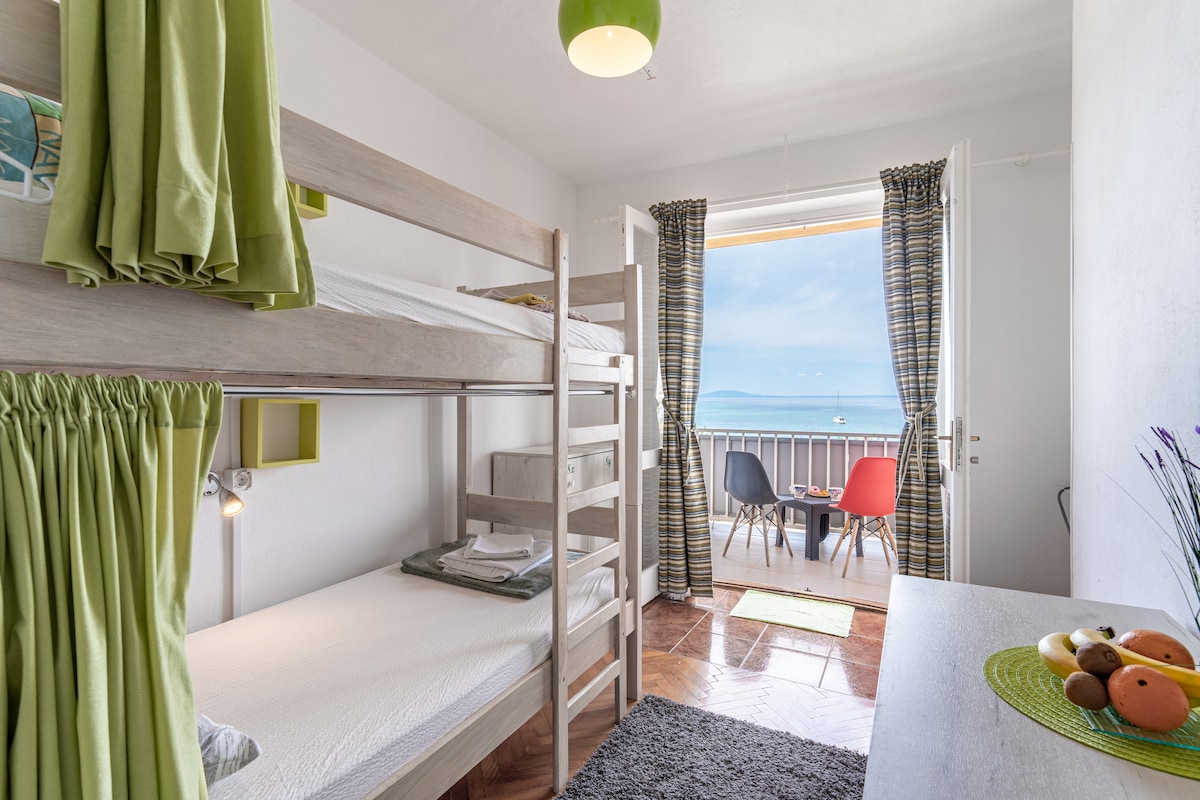 A bright room is shown with a bunk bed featuring soft linens. A door opens to a balcony, offering a view of the sea. A small dining table with colorful chairs sits on the balcony, complemented by a rug and a simple table arrangement inside.