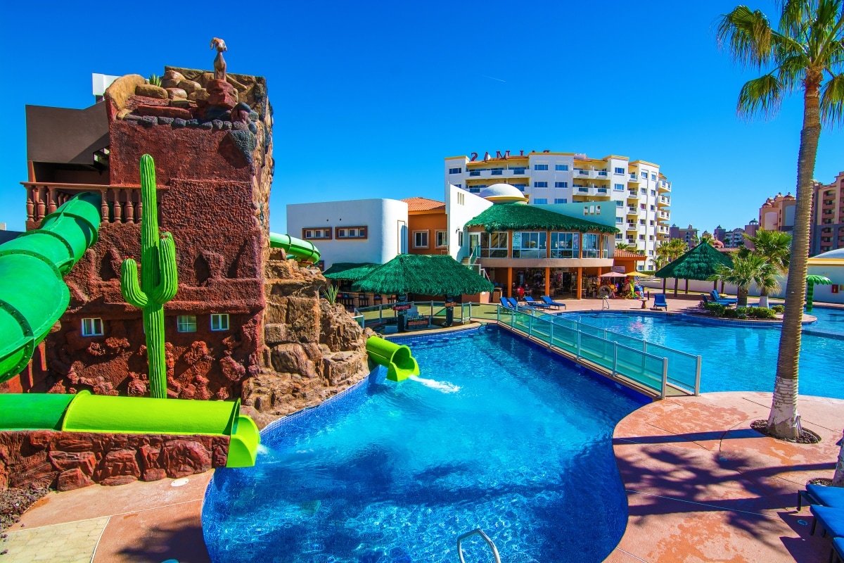 A large swimming pool features bright blue water, surrounded by palm trees and shaded seating areas. Colorful green water slides emerge from a rock formation, adding a playful element to the serene setting. The backdrop showcases a multi-story building and additional resort facilities.