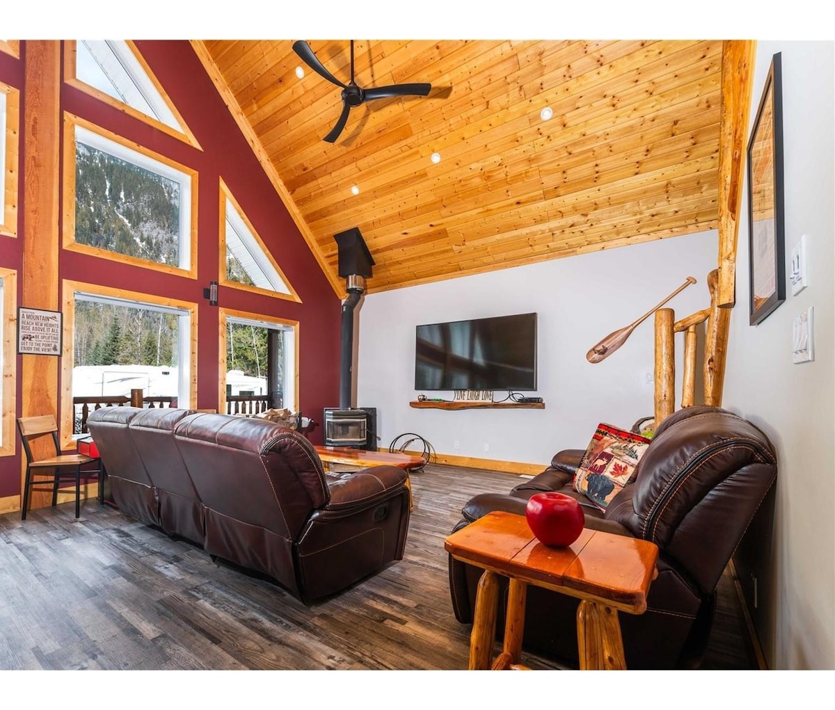 A spacious living area is characterized by a high wooden ceiling and large windows offering views of the outdoors. Comfortable leather seating is arranged around a coffee table, with a wood-burning stove and wall-mounted TV as focal points.