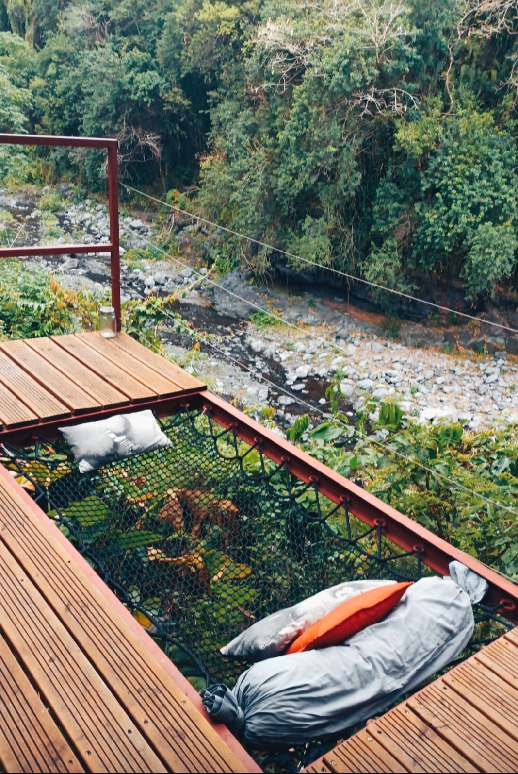A suspended netting area is set on a wooden deck, adorned with soft pillows in various colors. Below, a tranquil river flows gently through lush greenery, offering a serene connection to nature.