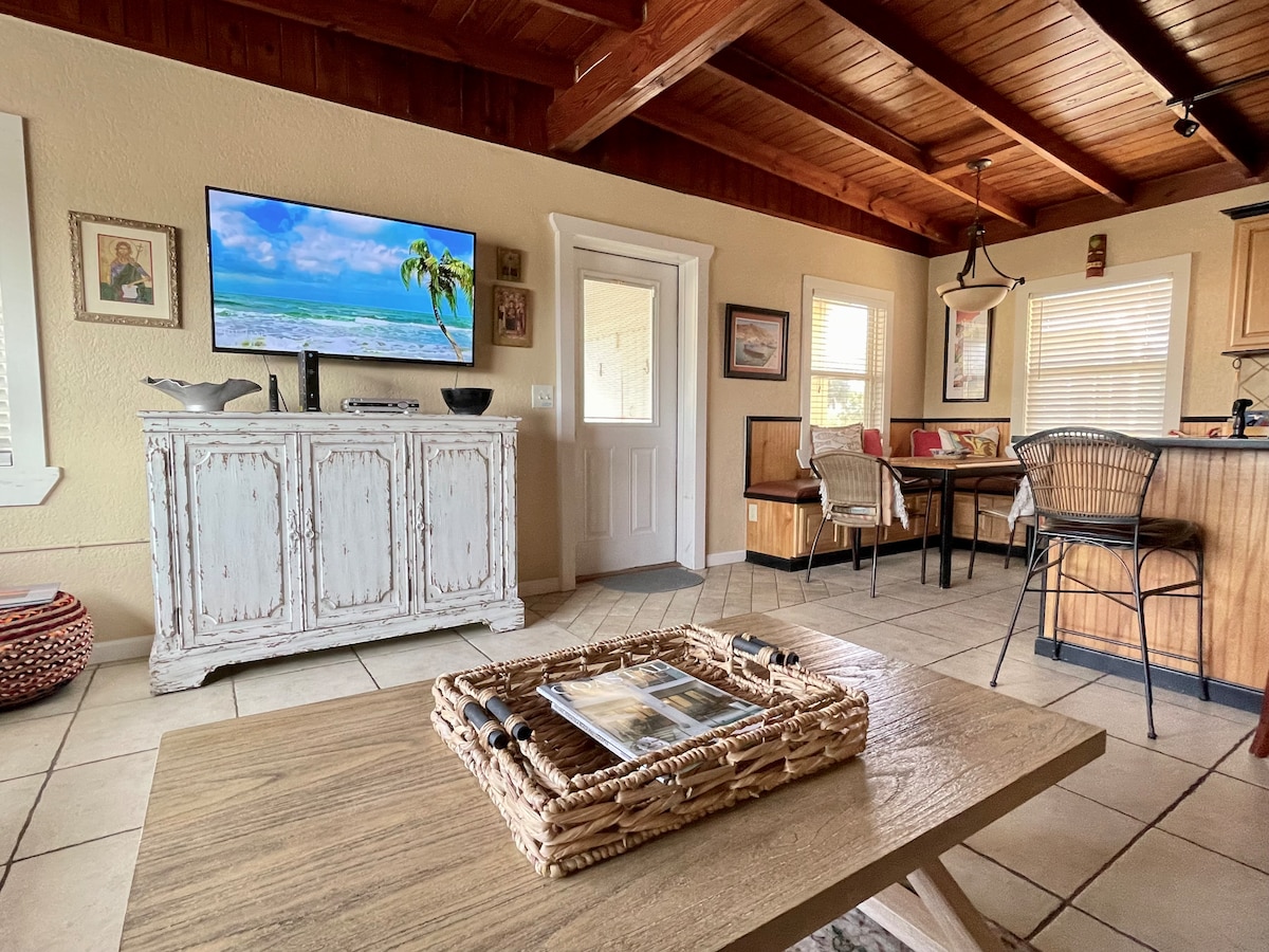 An inviting open living area features a rustic white cabinet with a television mounted above, displaying ocean scenery. A wooden coffee table holds magazines and a woven basket. Nearby, a dining space with seating for four and a cozy nook is visible.