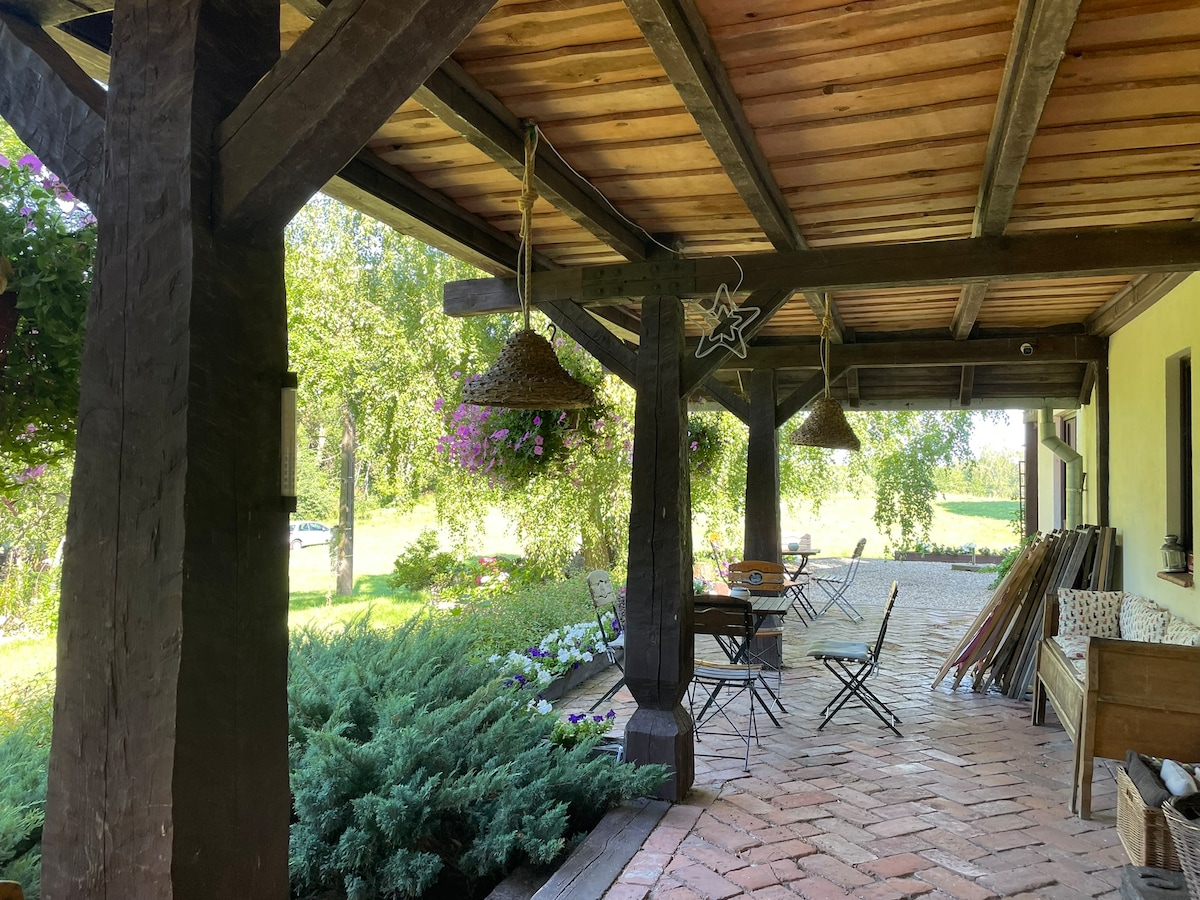 An outdoor terrace area features wooden beams supporting a shaded roof. Two hanging flower baskets add color, while a set of metal chairs and tables are arranged for dining. Lush greenery and blooming flowers surround the space, providing a serene atmosphere.