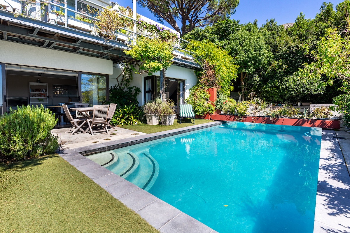 A vibrant outdoor area features a sparkling swimming pool surrounded by well-maintained gardens. Comfortable seating is arranged near the pool, offering a relaxing space to enjoy the lush surroundings, with greenery framing the area and enhancing the tranquil atmosphere.