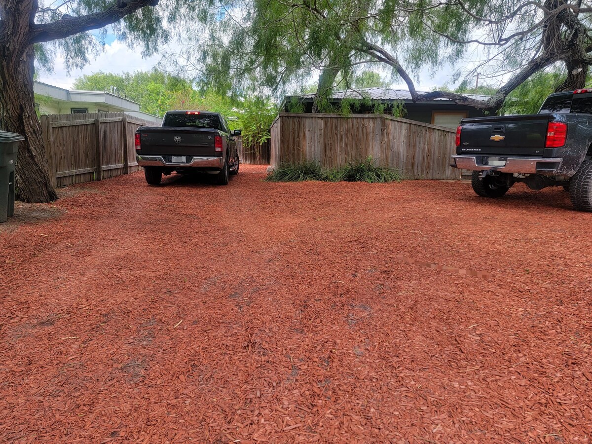 An outdoor parking area is depicted, featuring a gravel surface covered in red mulch. Two black pickup trucks are parked side by side, surrounded by wooden fencing and mature mesquite trees providing shade. The layout appears spacious, accommodating additional vehicles.