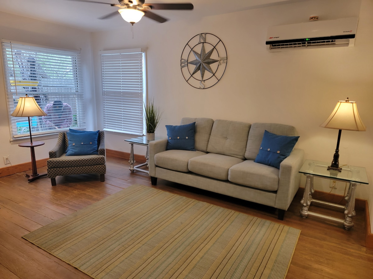 The living room features a light gray sofa complemented by blue accent pillows, flanked by two stylish lamps. A decorative wall clock with a star motif is mounted above the couch, and large windows allow natural light to fill the space, illuminating the reclaimed wood floor.