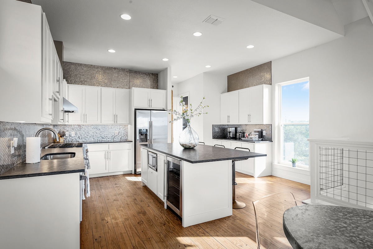 A modern kitchen features white cabinetry and a large island with bar seating. A stainless-steel refrigerator and sleek appliances are present. Natural light enters through a window, illuminating the polished wooden floor and highlighting the contemporary design.