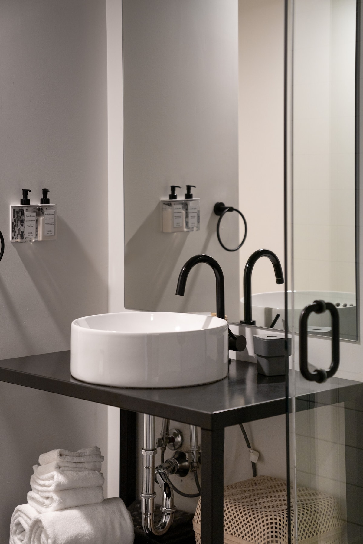 A modern bathroom features a white vessel sink resting on a sleek black vanity. Stylish fixtures in matte black contrast with the minimalist decor. Soft towels are neatly stacked nearby, while decorative elements add subtle warmth to the space.